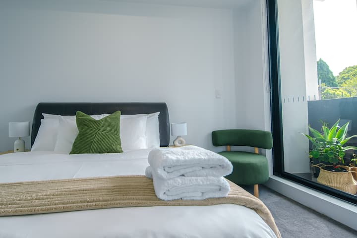 Cama tamaño queen con sábanas blancas limpias y una manta suave con textura. Las mesitas de noche con lámparas agregan calidez, mientras que la luz natural fluye desde las puertas del balcón.