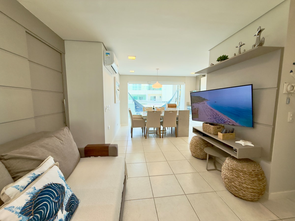 Élégant salon avec canapé beige décoré de coussins bleus, panneau TV avec bibliothèque et télévision à écran plat, sol en porcelaine et éclairage naturel; détendez-vous en regardant la télévision et profitez de l'intégration avec la salle à manger!