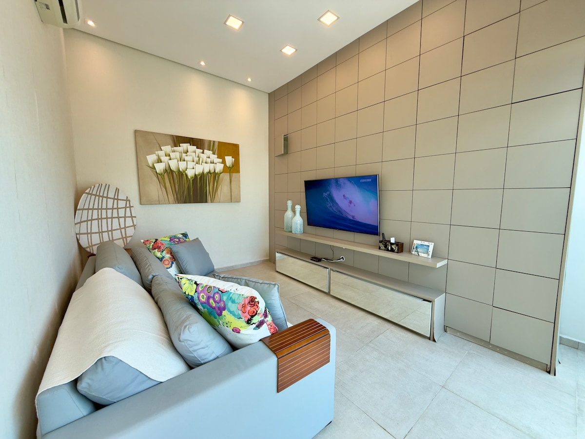 Salon moderne avec canapé gris et coussins colorés, panneau en bois avec téléviseur à écran plat, cadre floral et éclairage intégré; détendez-vous confortablement en regardant vos émissions préférées et en profitant du décor élégant!