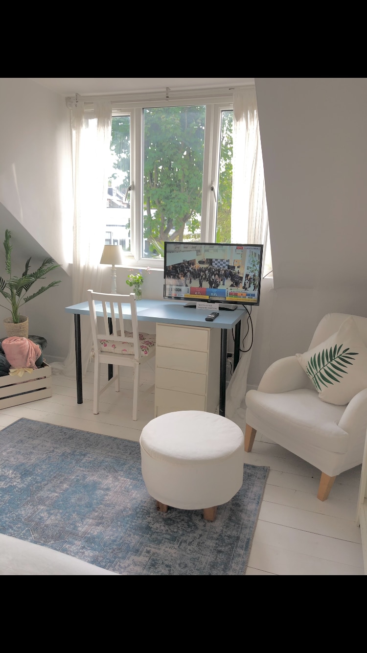 A well-lit workspace features a small desk with a computer monitor and a lamp, alongside a white chair and an ottoman. A decorative plant adds a touch of greenery, while the window reveals a view of trees outside, enhancing the light and airy ambiance.