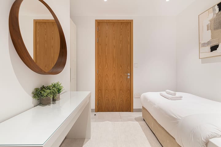 A bright single bedroom with a neatly made bed, bedside table, and a vanity topped with a round mirror and decorative accents. The clean design and calm atmosphere make it a comfortable space to rest or recharge.