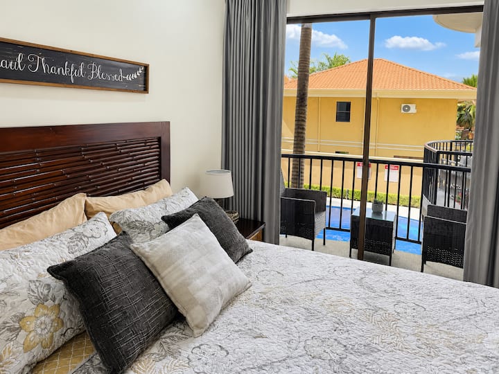 Comfortable bedroom with layered pillows, wood headboard, soft neutral bedding, and large sliding doors opening to a private balcony with seating and pool views.