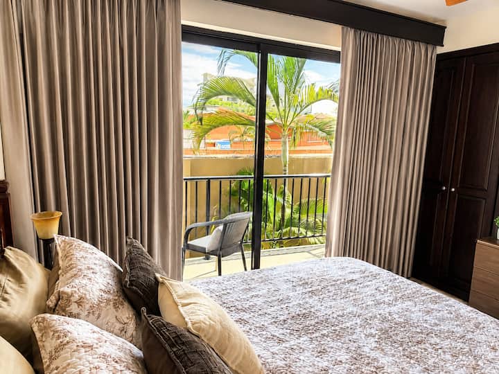 This inviting bedroom opens onto a private balcony through large sliding glass doors. Floor-to-ceiling curtains provide privacy, while tropical views and outdoor seating create a bright, relaxing place to begin or end the day.
