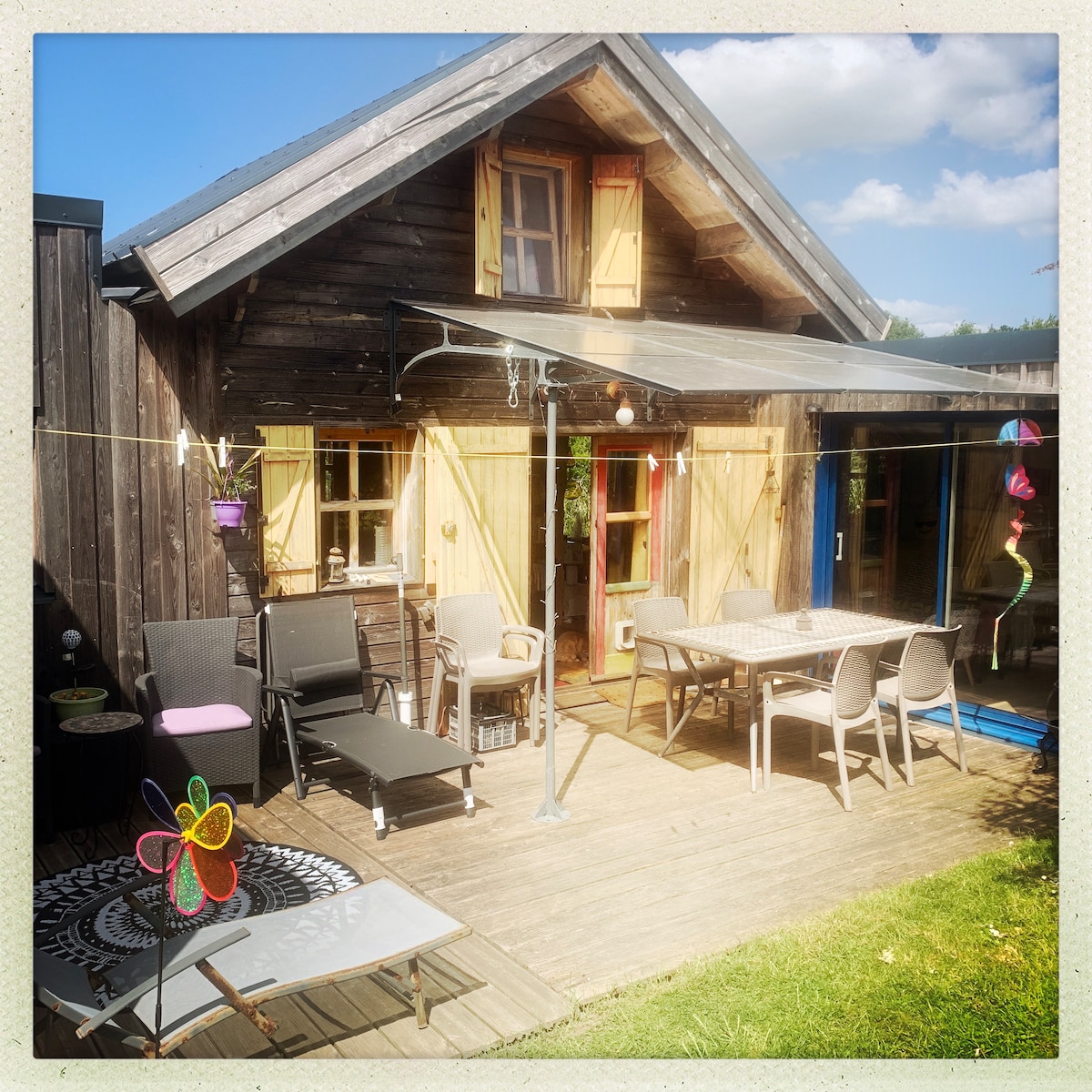 An outdoor terrace is featured, surrounded by wooden cabin walls. A spacious dining table is visible, accompanied by several seating options including lounge chairs. A partially shaded area provides comfort, with colorful decorations adding a playful touch. Sunlight illuminates the scene, creating a relaxed atmosphere.
