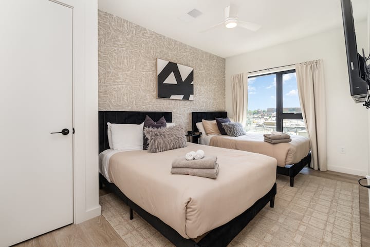 Bright guest bedroom with two plush beds, soft neutral bedding, and textured accent wall. Black headboards add contrast, while large window fills the room with natural light and city views. Clean, modern, and hotel-style.