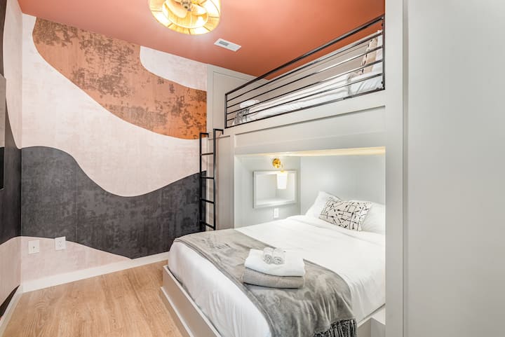 Modern bunk-style bedroom with custom loft bed, built-in ladder, and bold mural accent wall in warm terracotta and charcoal tones. Soft lighting, clean lines, and layered textures create a cozy yet design-forward space.