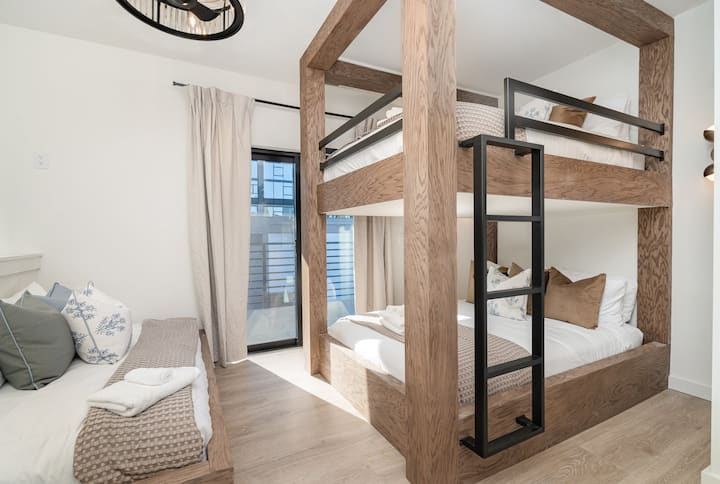 Modern bunk room with custom wood-built beds, black metal ladder, and clean white walls. Natural light pours in from the balcony door, while layered bedding and neutral textures create a cozy, upscale retreat feel.