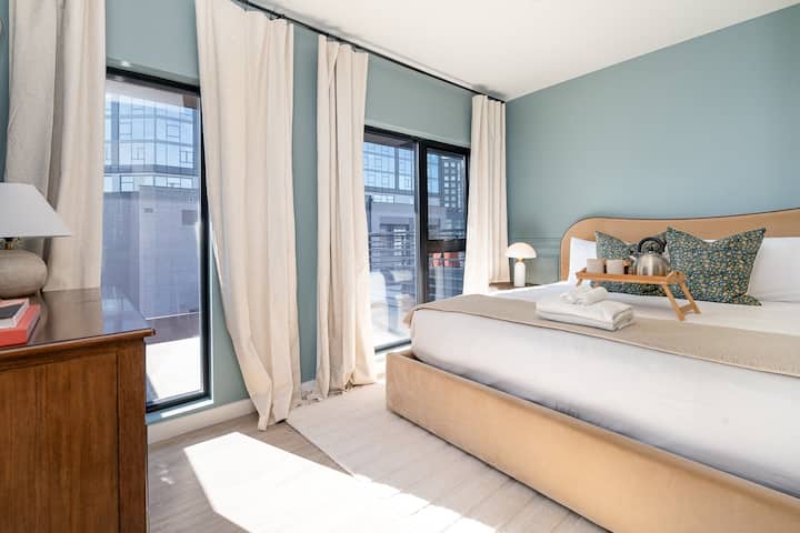 Bright, calming bedroom with soft blue walls, floor-to-ceiling windows, and sheer curtains that fill the space with natural light. Upholstered bed, layered linens, and warm wood accents create a relaxed, boutique-hotel feel.