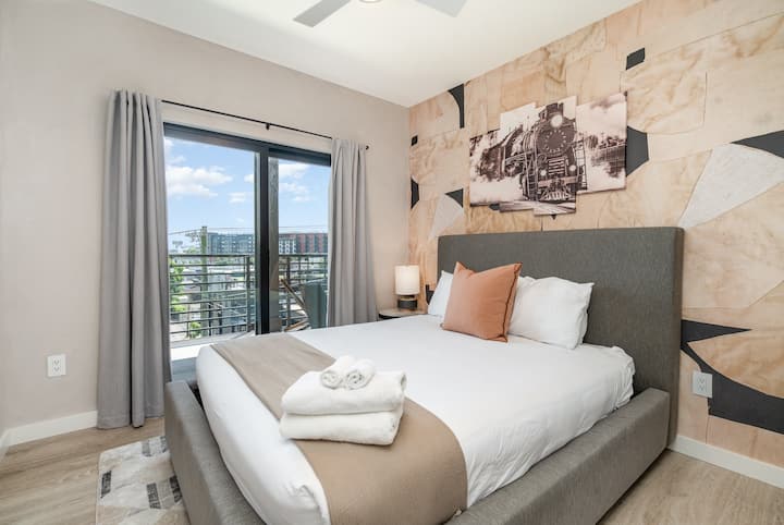 Modern bedroom with upholstered gray bed, crisp white linens, and warm accent pillow. Feature wall with abstract wood tones and train artwork adds character, while sliding glass doors open to a private balcony with city views.
