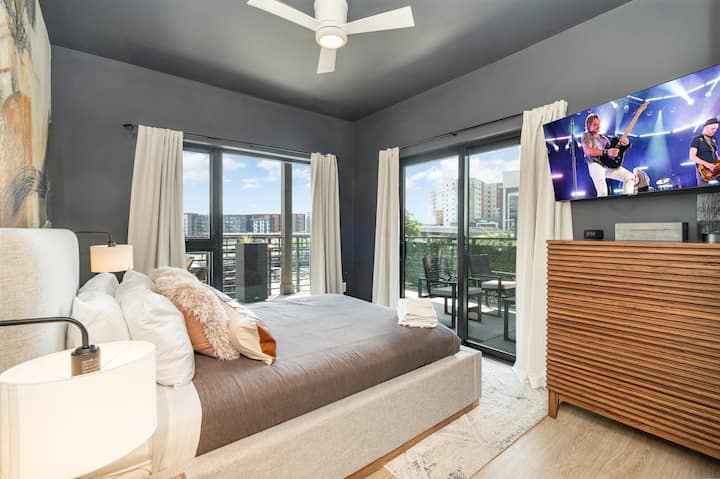 Moody modern bedroom with charcoal walls, plush upholstered bed, layered neutral bedding, and warm accent pillows. Sliding glass doors open to a private balcony, while a mounted TV and slatted wood dresser add clean, contemporary style.