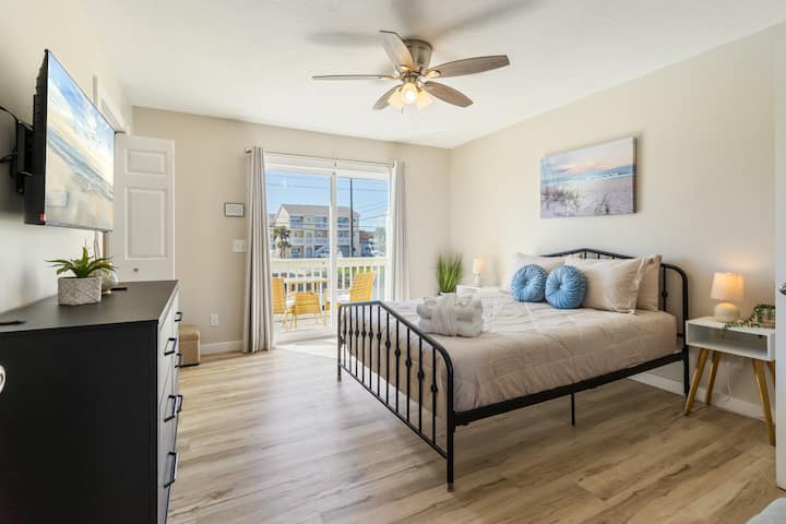 Útulná, slnkom zaliata spálňa s pohodlnou manželskou posteľou Queen, dekoráciami s plážovou tematikou a televízorom namontovaným na stene. Vyznačuje sa mäkkými neutrálnymi tónmi, štýlovými nočnými stolíkmi s teplým osvetlením a posuvnými sklenenými dverami vedúcimi na súkromný balkón s vonkajším posedením.