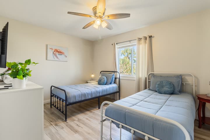 Svetlá a útulná spálňa s dvoma samostatnými posteľami s mäkkou modrou posteľnou bielizňou, stropným ventilátorom a svetlým dekorom. Zahŕňa komodu s televízorom, prirodzené svetlo z okna a jednoduché vybavenie ideálne na relaxačný pobyt na pláži.