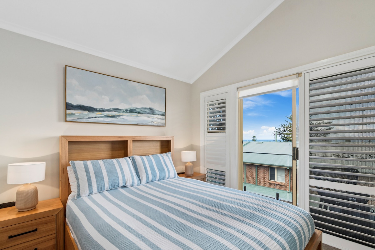 Recharge in the primary bedroom, where a queen bed dressed in crisp linens, ocean glimpses, and soft blue accents reflect the calm rhythm of the coast.