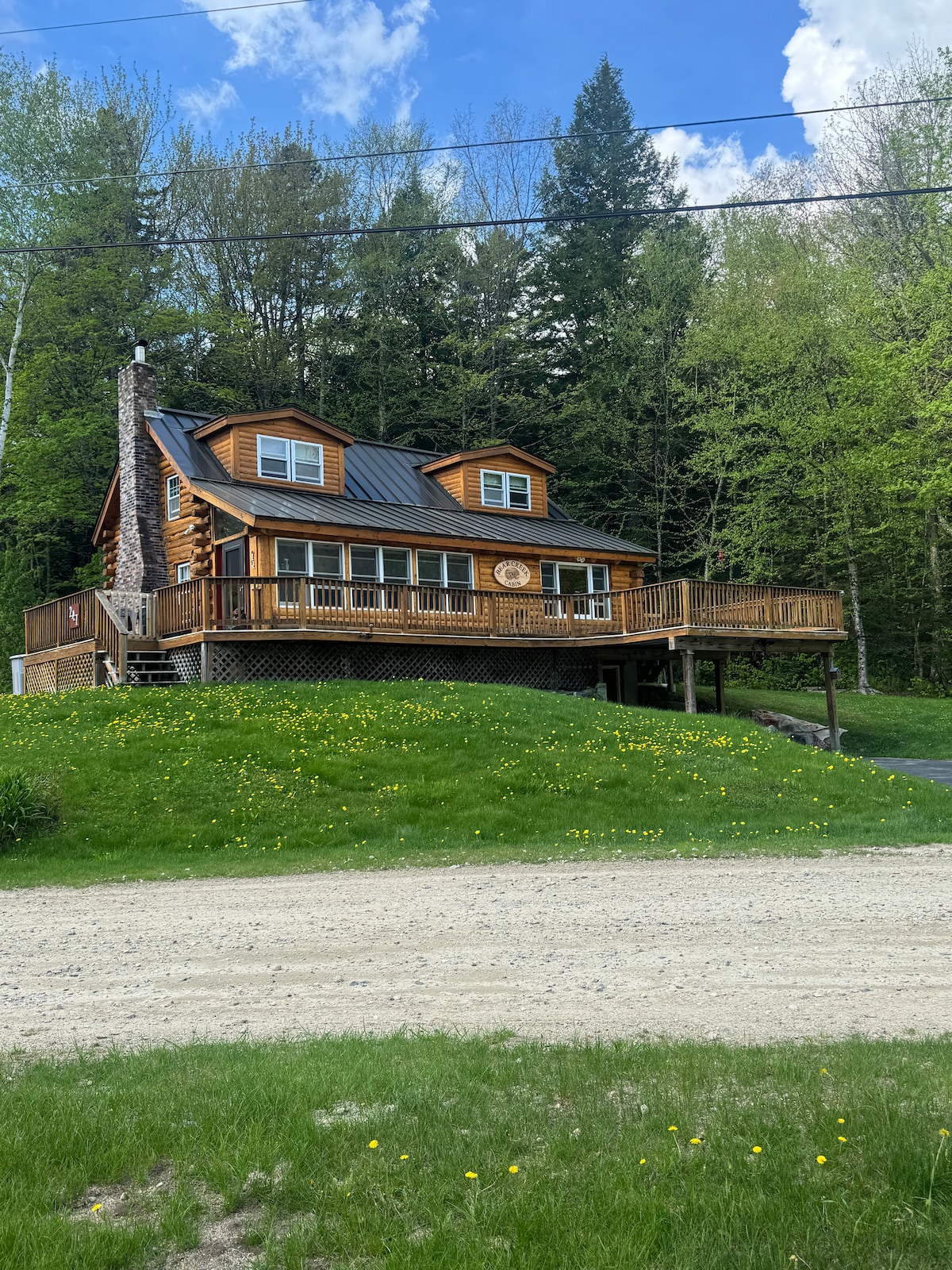 A log cabin is situated on a grassy knoll, surrounded by green foliage. The structure features a large front deck with wooden railings and a clock mounted on the facade. The roof is clad in dark metal, and the driveway is visible leading to the cabin.