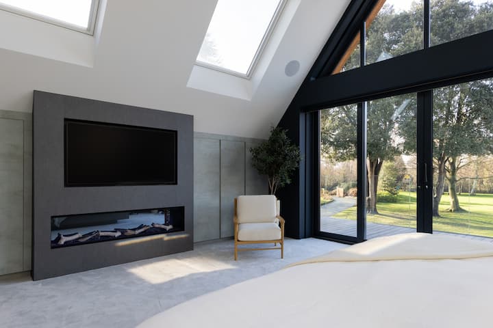 Stylish master bedroom  with vaulted ceilings, skylights, and large windows offering garden views. Featuring a modern fireplace and comfortable seating, it’s a bright and relaxing space.