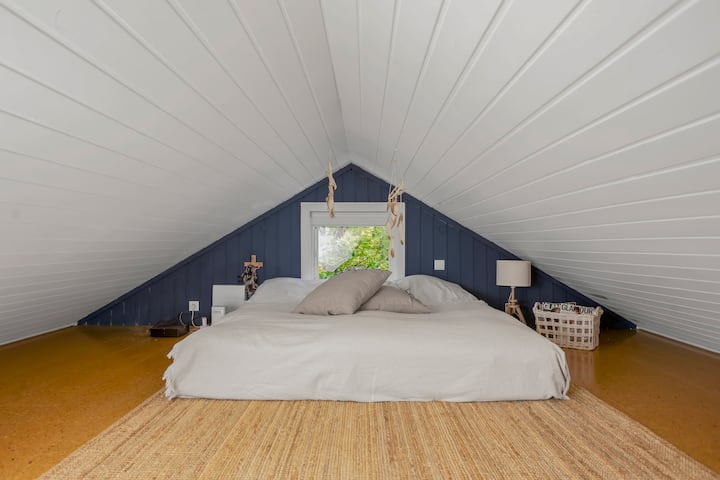 Уютно лофт пространство с ниско легло под наклонен бял таван. Меко спално бельо в неутрални тонове, топли дървени подове и прозорец, който внася естествена светлина. Простият декор създава чист и комфортен вид.