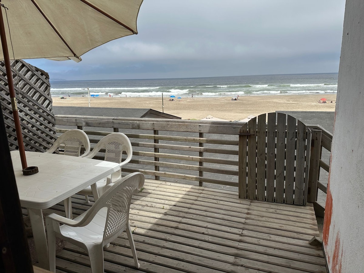 A wooden terrace is shown with a white table and several white chairs, providing a comfortable outdoor dining space. The ocean is visible in the background, with a sandy beach and scattered beachgoers enhancing the coastal atmosphere.