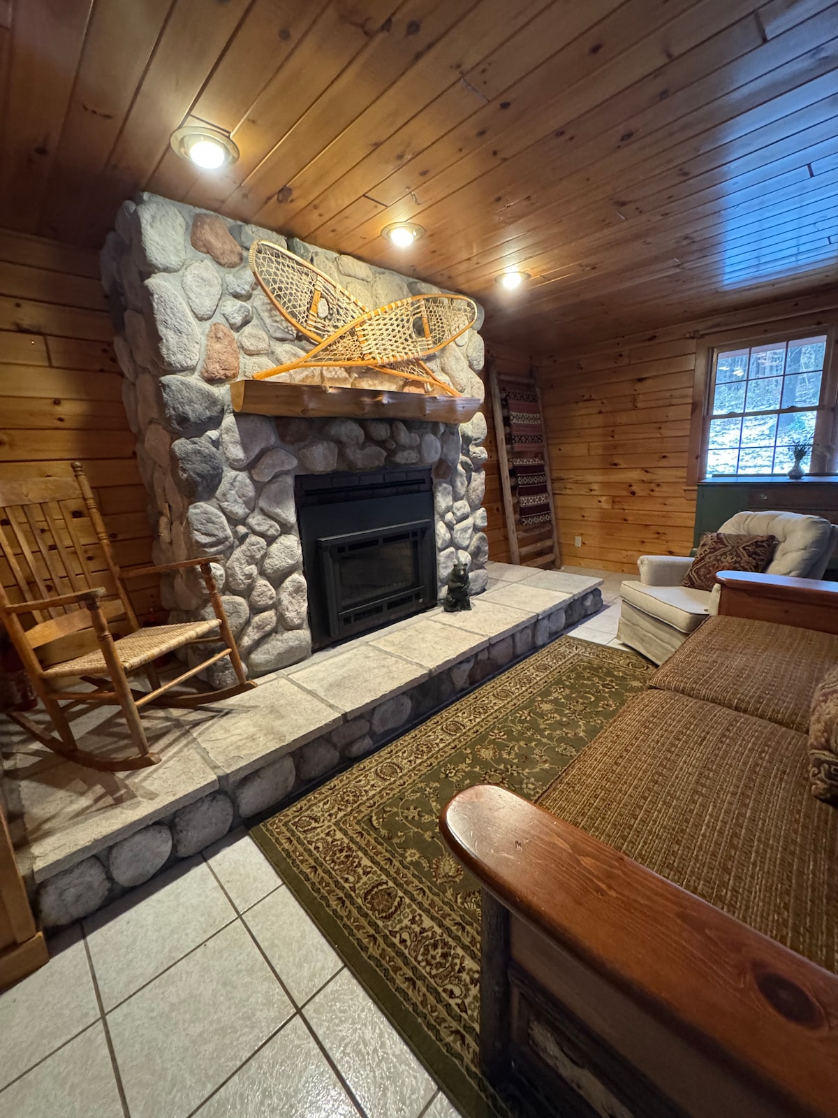 Bright Adirondack-style living room with warm wood accents, comfortable seating, fireplace, and large windows overlooking the 30-acre property. Newly renovated space designed for relaxing after exploring the private cabin’s trails and waterfall.