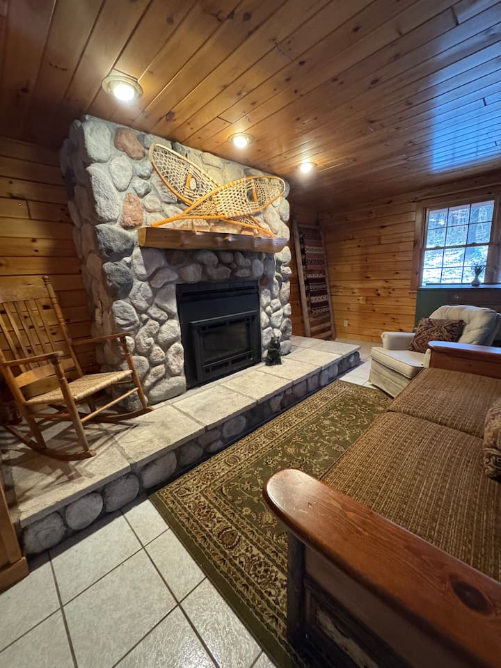 Bright Adirondack-style living room with warm wood accents, comfortable seating, fireplace, and large windows overlooking the 30-acre property. Newly renovated space designed for relaxing after exploring the private cabin’s trails and waterfall.
