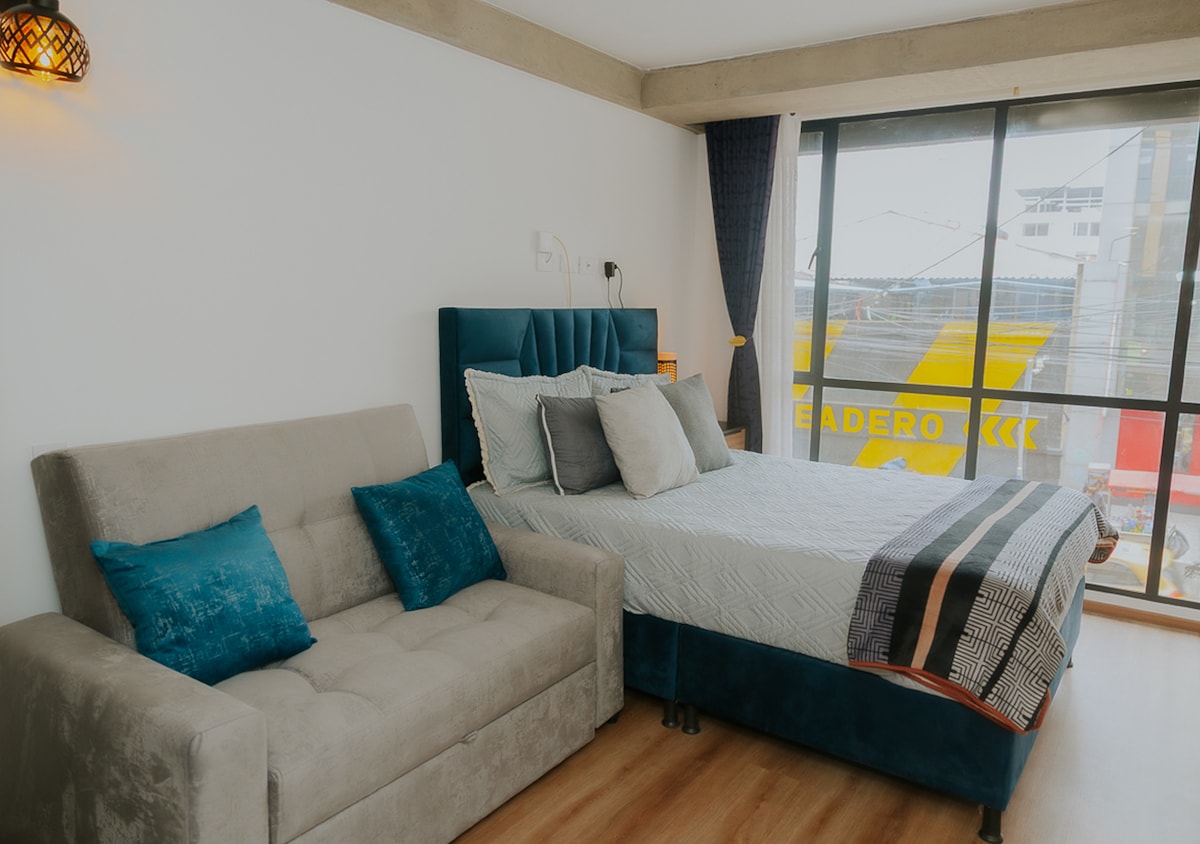 Moderno y acogedor estudio con cama doble, sofá gris y ventanales que llenan el espacio de luz natural. Decorado con tonos azul y gris, ofrece un ambiente elegante y confortable ideal para descansar o trabajar.