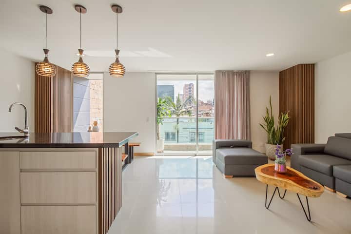 Ambiente abierto y luminoso con ventanales de piso a techo, sala moderna, detalles en madera y plantas naturales. Un espacio que invita a relajarse, compartir y disfrutar de luz, estilo y comodidad.