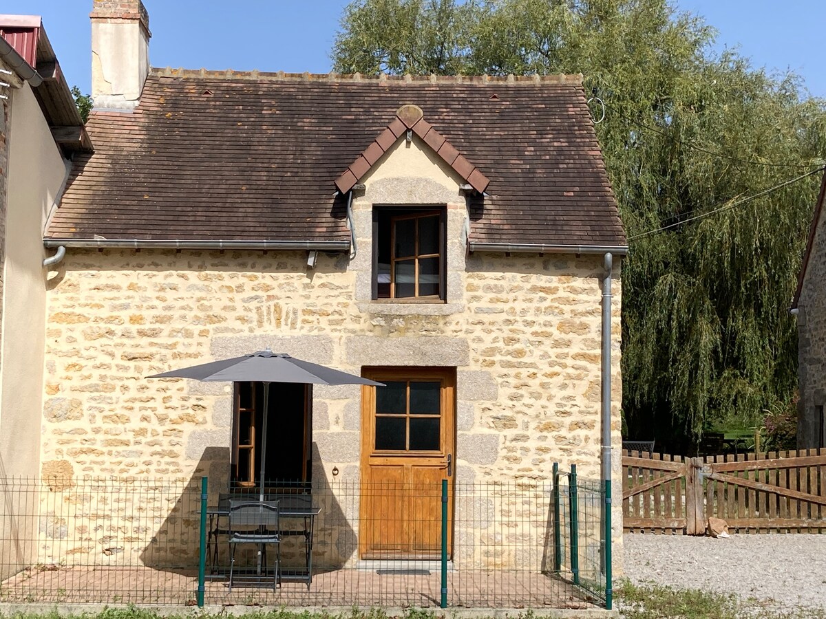 A charming countryside house is presented, featuring a stone facade and a sloped roof with brown shingles. In front, a small outdoor seating area is arranged under a grey patio umbrella, complemented by a wooden door and two windows that allow natural light.