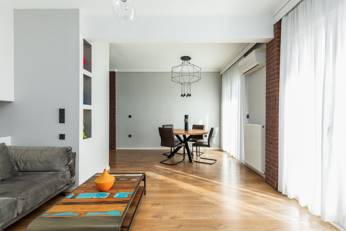 Svetlý a otvorený obytný priestor s modernými sivými tónmi, drevenými podlahami a farebným konferenčným stolíkom, ktorý plynule prechádza do jedálenského kúta – ideálny na oddych alebo zábavu.