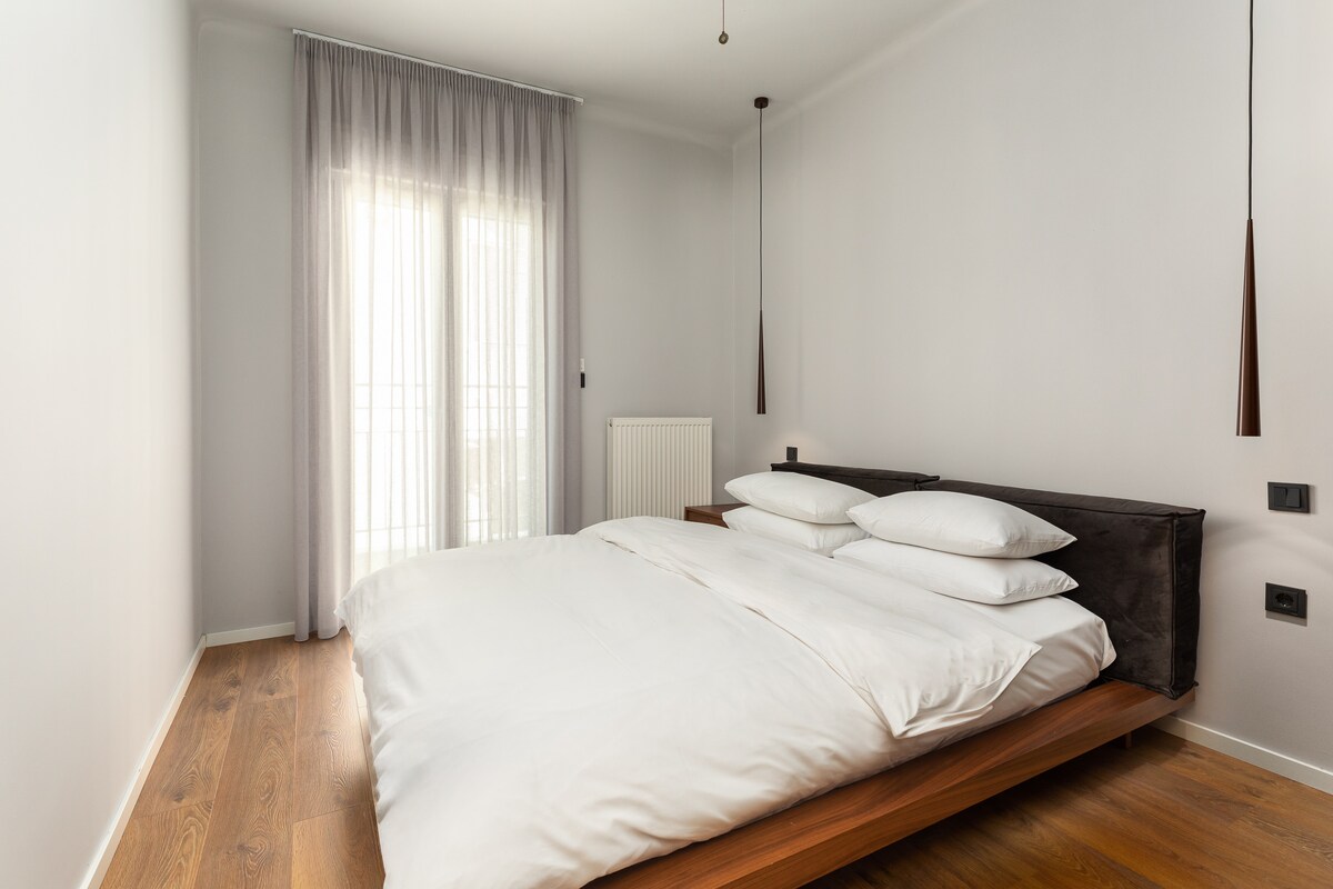 Svetlá a minimalistická spálňa s pohodlnou manželskou posteľou, priehľadnými závesmi a teplými drevenými podlahami, ktoré vytvárajú pokojné a vzdušné útočisko.