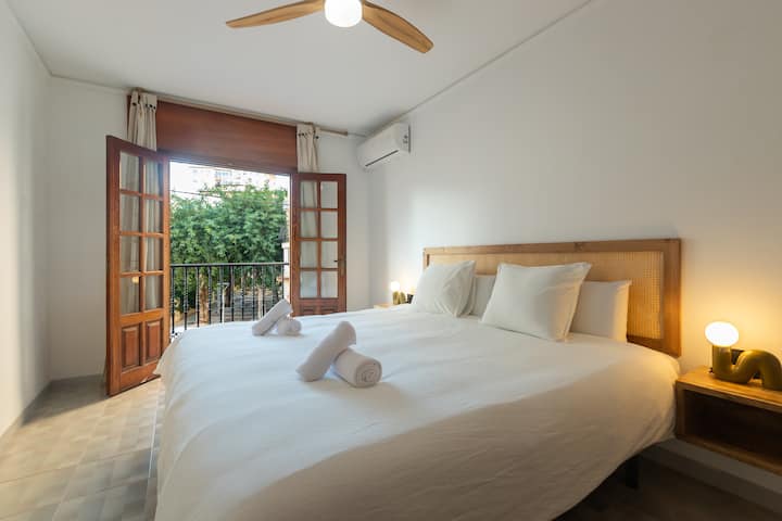 Dormitorio amplio y muy luminoso con cama doble, cabecero de madera y ropa de cama blanca. Ventilador de techo y aire acondicionado. Puertas francesas de madera que se abren al balcón con barandilla de forja y vistas a los árboles.