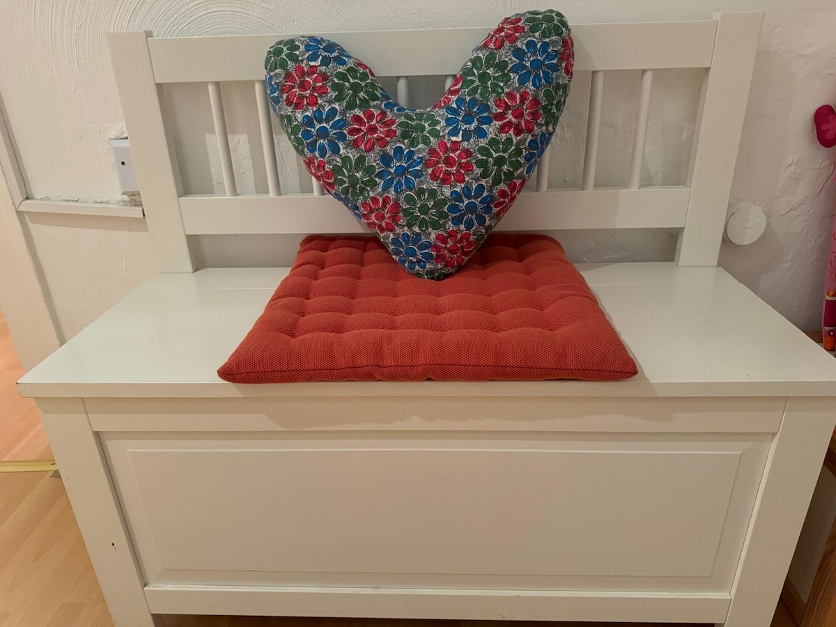 A wooden bench is adorned with a colorful heart-shaped cushion and a bright orange seat pad. The bench features a classic slatted design, providing a charming and functional seating area. The simple decor complements the surrounding space.