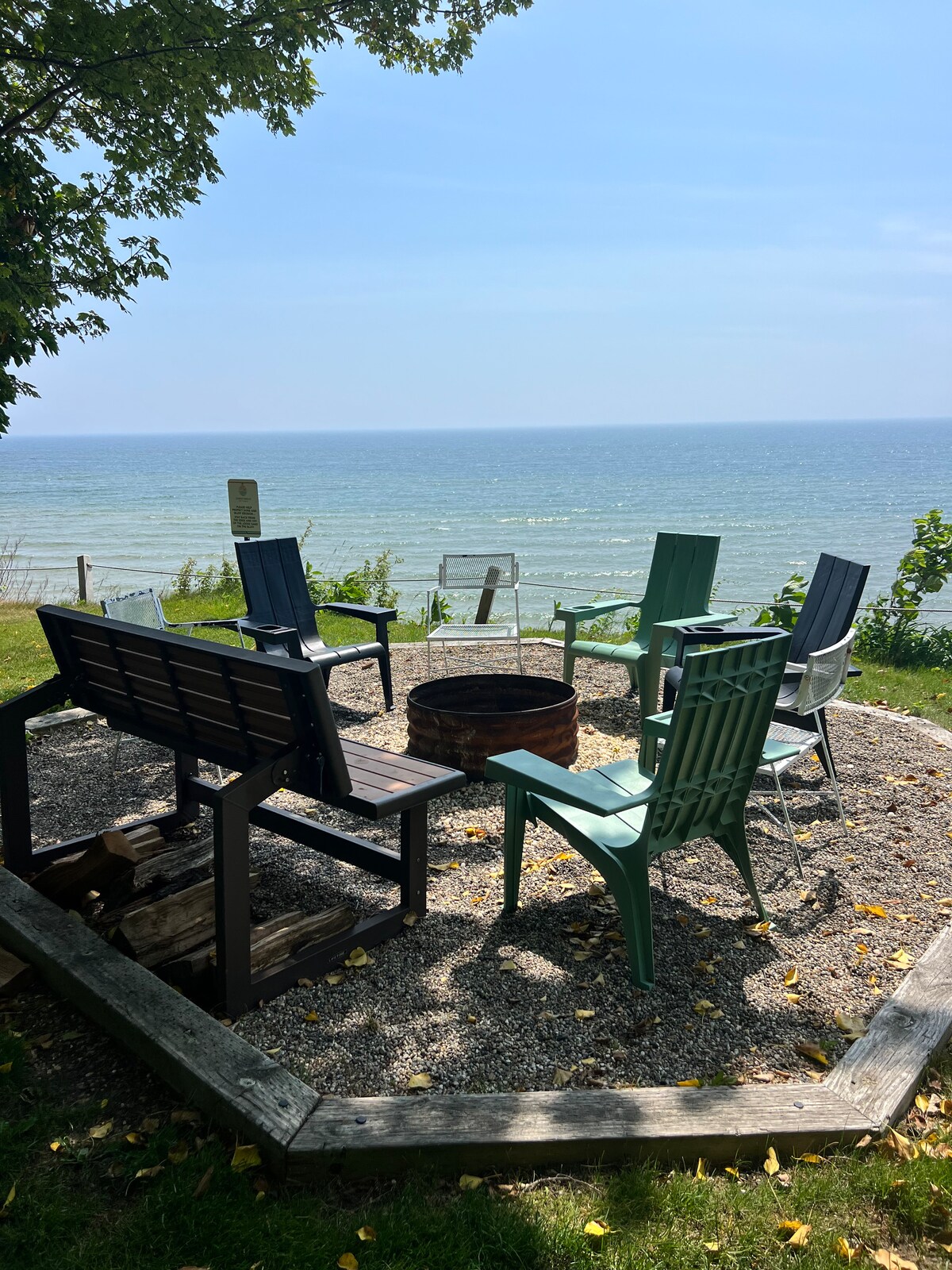 A fire pit area is surrounded by six chairs in varied colors, creating an inviting space for gatherings. The pit is set in a circular gravel area, offering views of Lake Michigan, with lush greenery framing the background.