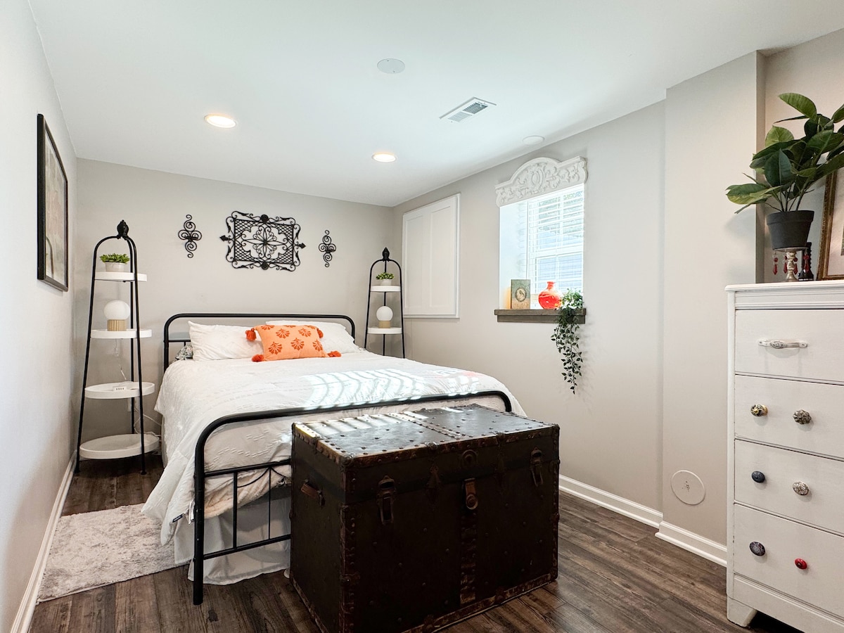 Dormitorio luminoso y acogedor con cama de estructura metálica, ropa de cama blanca y decoración cálida. Cuenta con estantes de cabecera a juego con lámparas, un baúl vintage, arte mural y luz natural para un relajante refugio.