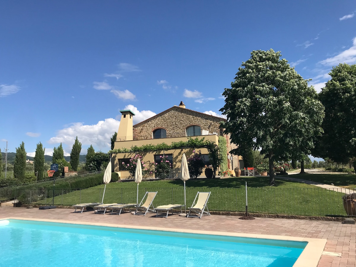 A traditional stone farmhouse is highlighted against a clear blue sky, surrounded by lush greenery. The large swimming pool, with lounge chairs and umbrellas, is positioned in the foreground, inviting relaxation amidst the serene vineyard landscape.