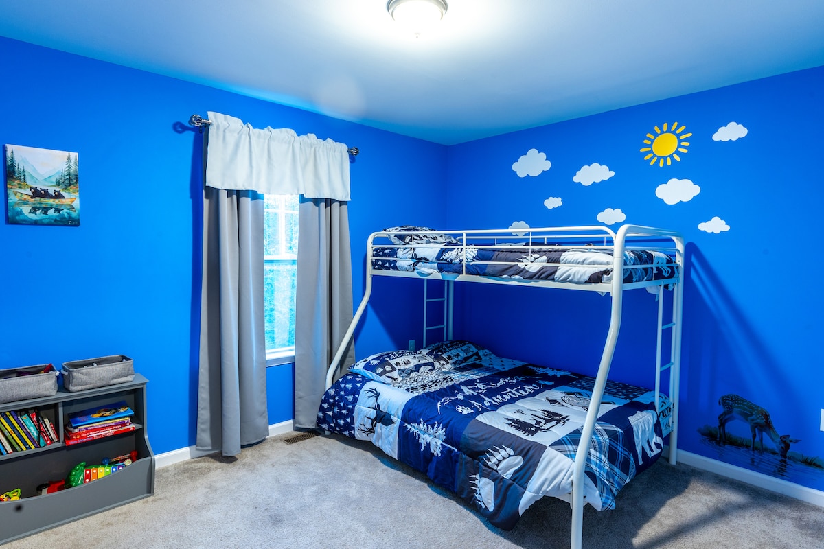 Chumba cha kulala chenye mwanga na kinachofaa watoto chenye vitanda viwili na kona ya starehe iliyojaa vitabu na midoli.