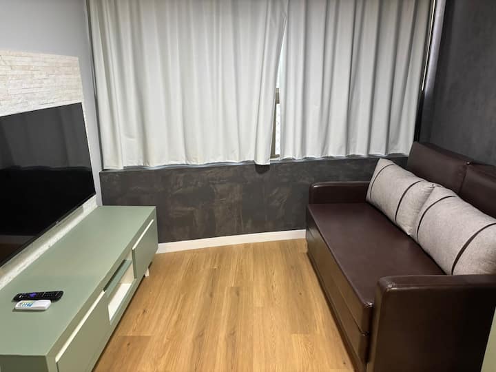 Acogedora sala de estar con cómodo sofá cama, TV inteligente y aire acondicionado. Ambiente bien iluminado con cortinas opacas, ideal para relajarse o alojar a un huésped adicional con comodidad y practicidad.