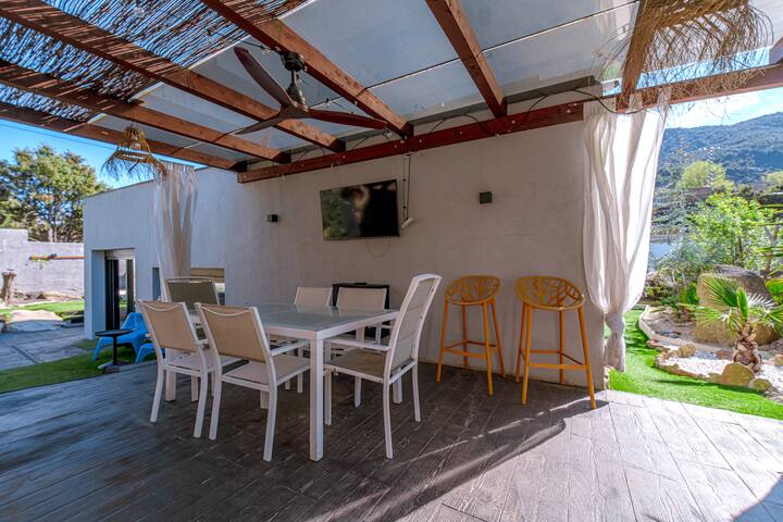 Comedor de verano bajo la pérgola: TV exterior, altavoces y cortinas para la sombra.