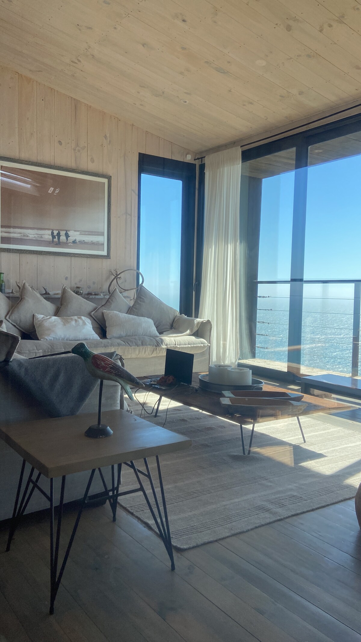 A bright living area features a comfortable sofa adorned with several cushions. Large glass doors open to a stunning view of the ocean, allowing natural light to fill the space. A small coffee table and decorative elements complete the cozy atmosphere.