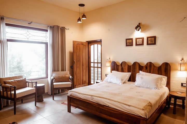 Rustic luxury defined. A solid wood king bed with a unique wavy headboard anchors the room. The large glass window floods the space with natural light, highlighting the "breathing" lime plaster walls.