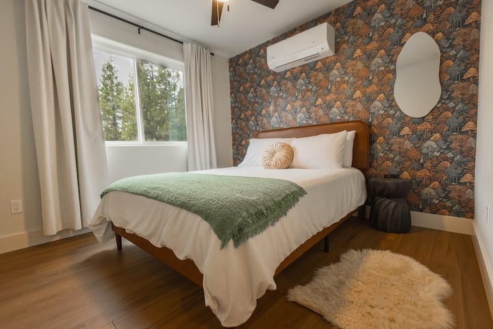 Downstairs bedroom features queen bed, comfortable bedding, extra pillows, blanket, warm lighting, 2 nightstands, mini split air conditioner / heater, electric wall heater, unique mirror, stylish wallpaper, and full bathroom next door in hallway.