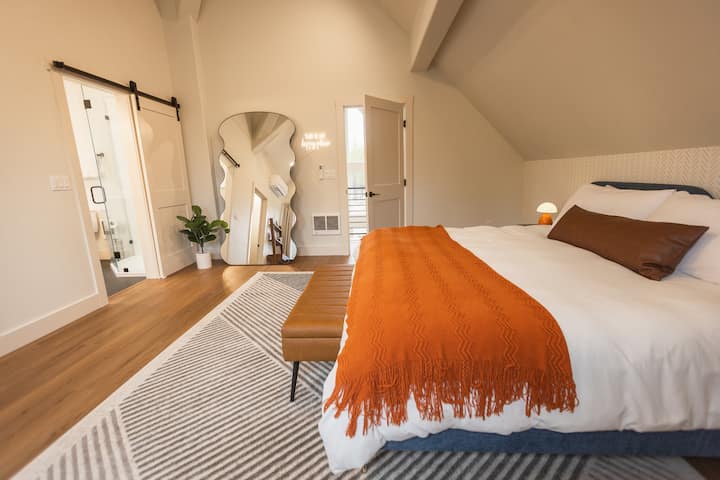 Upstairs bedroom features king bed, comfortable bedding, extra pillows, blanket, clothes storage (with hangers), warm lighting, mini split air conditioner / heater, electric wall heater, full length mirror, LED sign, and private full bathroom.