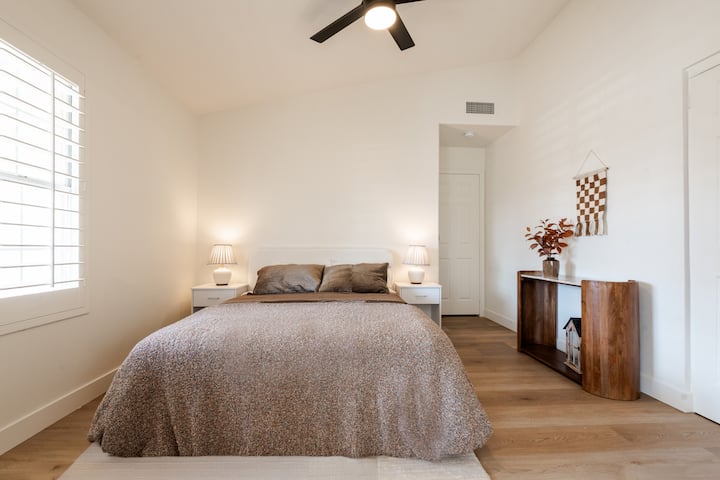 Quarto iluminado e minimalista com uma cama confortável, iluminação aconchegante e decoração neutra moderna. A grande janela traz luz natural suave, criando um espaço tranquilo para descansar e recarregar as energias.
