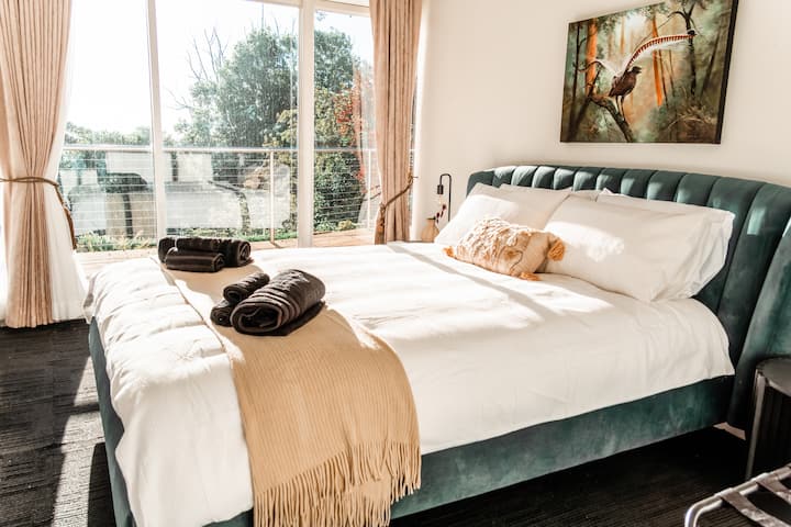 A jewel-toned queen room with a forest green velvet bedhead, Australian wildlife art, direct bathroom access, and treetop garden views