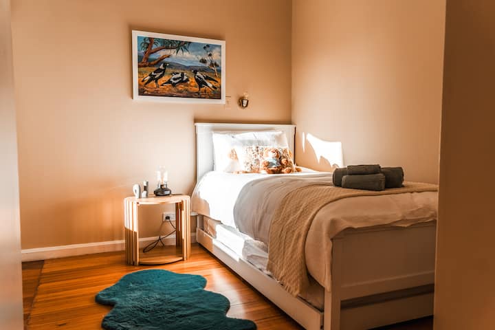 A warm terracotta single-and-trundle room with adult-quality mattresses, a magpie artwork, teal faux sheepskin rug, and treetop views