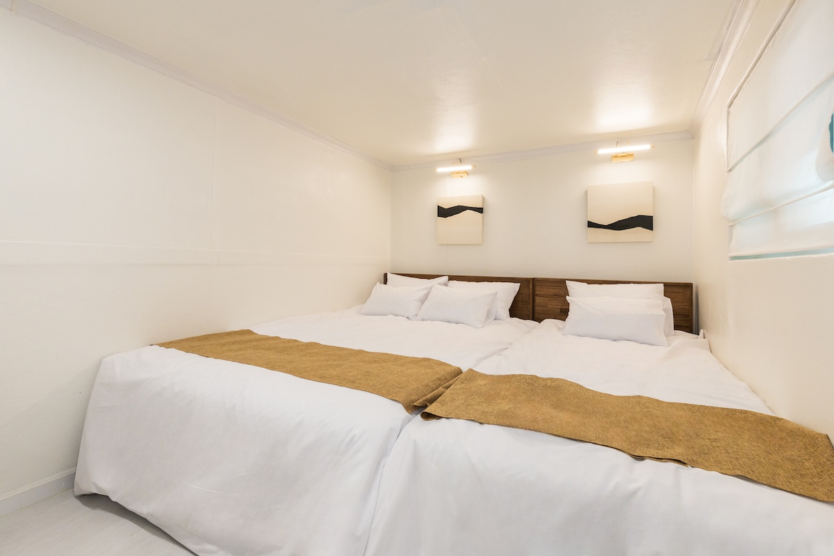 מיטת הקווין סייז והמיטה הרכה והנוחות ממוקמות זו לצד זו, ונראות כמו מיטה מרווחת, והן מעוטרות בצורה מסודרת במצעים נקיים ובשטיחים חומים. ראש המיטה מעץ מעניק תחושת יציבות, והקיר מעוטר בציורים אבסטרקטיים של קימורים שחורים ותאורה כדי להוסיף אווירה מתוחכמת. קירות נקיים, תקרות ותריסי חלונות משלימים חלל מואר ונקי, המעניק לכם את החמימות של מלון קוריאני מסורתי. המיטות מחוברות, כך שתוכלו להשתמש בהן עם ילדכם מבלי לדאוג למרווחים.