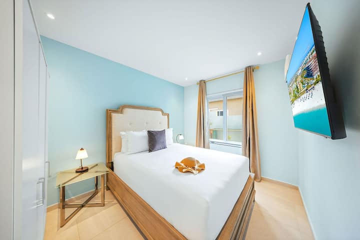 Plongez dans le lit confortable, regardez la télévision et détendez-vous dans la chambre lumineuse et sereine avec des murs bleus doux, un éclairage chaleureux et la lumière naturelle d'une grande fenêtre.