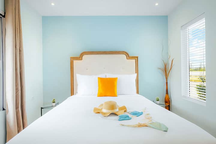 Profitez d'un lit confortable avec des draps blancs, une touche d'orange ensoleillée et des murs bleus lumineux, parfaits pour se réveiller rafraîchi et prêt pour des aventures sur l'île.