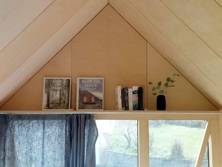 الجملون فوق السرير مع رف كتب مدمج. يتم توفير كتب الهندسة المعمارية والخيال للاطلاع عليها. يضيف سقف خشب البتولا الرقائقي المثلث الشكل الارتفاع والشخصية إلى الغرفة الصغيرة.