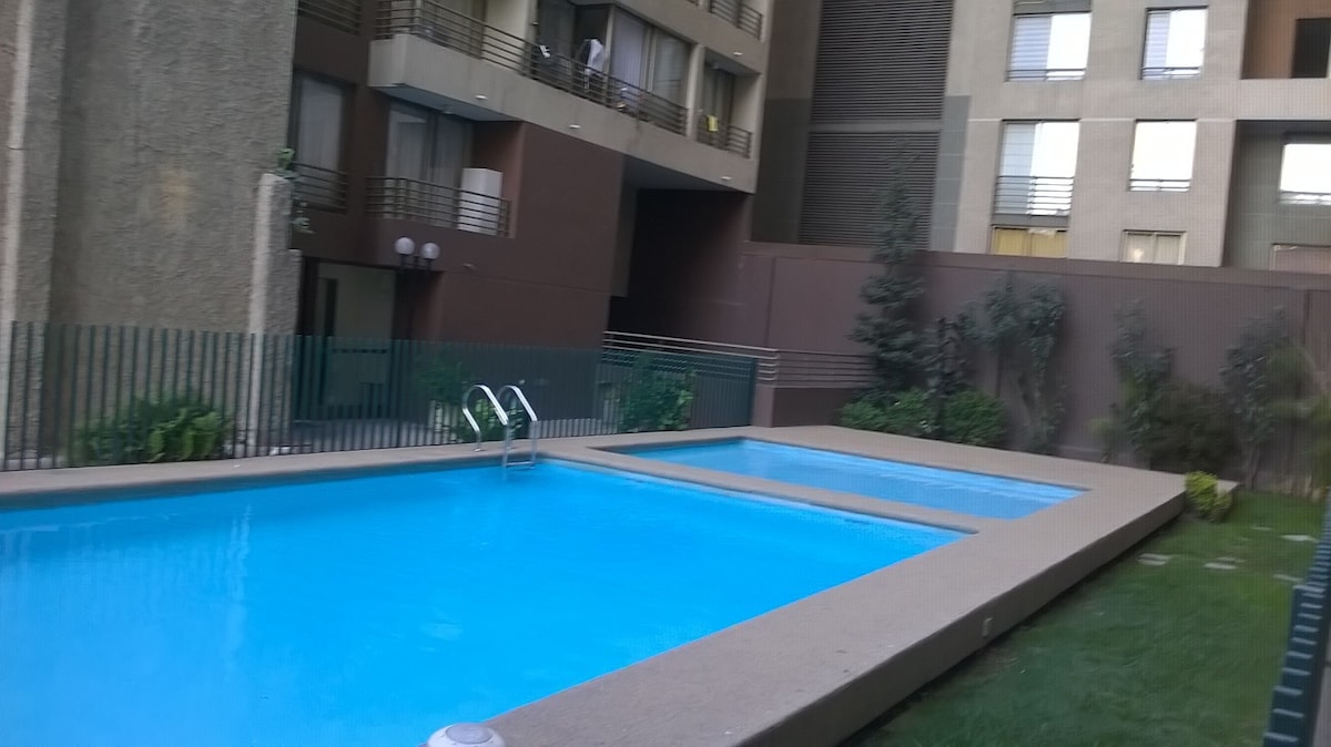 A well-maintained outdoor swimming pool area is visible, featuring two pools with clear blue water. Sun loungers are arranged around the pool, and green plants add a touch of nature. The area is surrounded by a low fence for privacy.