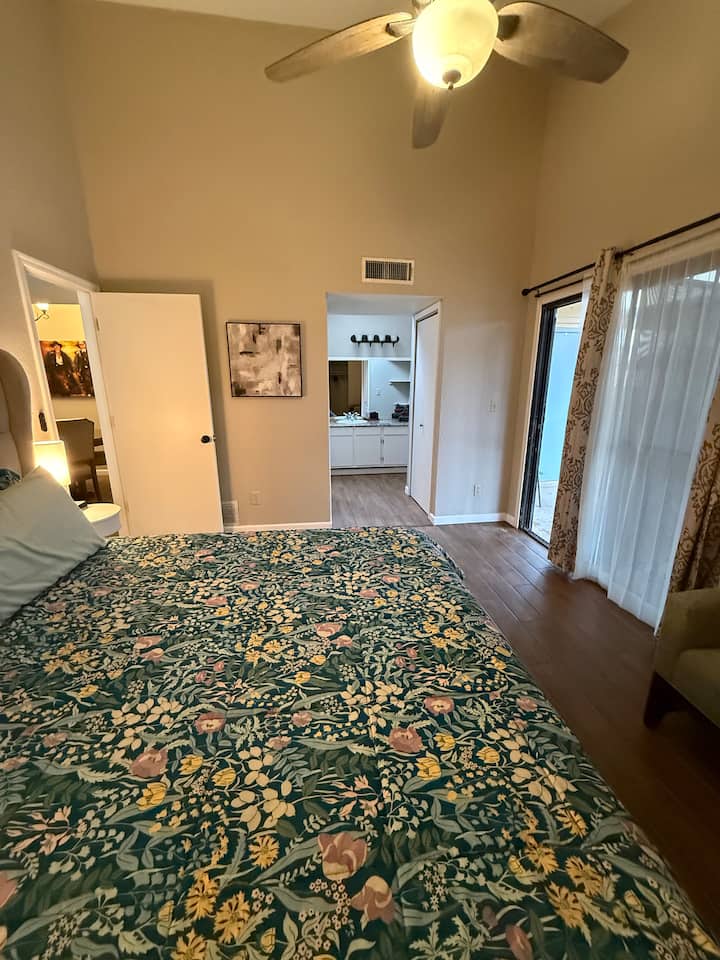 King size bed with lighted ceiling fan, ensuite bathroom, sliding door with direct back yard access, small seating couch, and television, along with a closet.