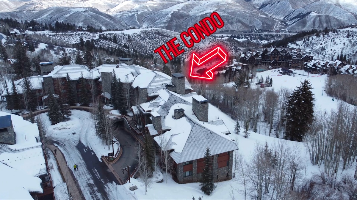 An aerial view showcases a snow-covered mountain landscape, highlighting a cluster of rustic buildings. The condo is indicated with a stylized arrow. Surrounding trees are blanketed in snow, contributing to the serene winter setting.